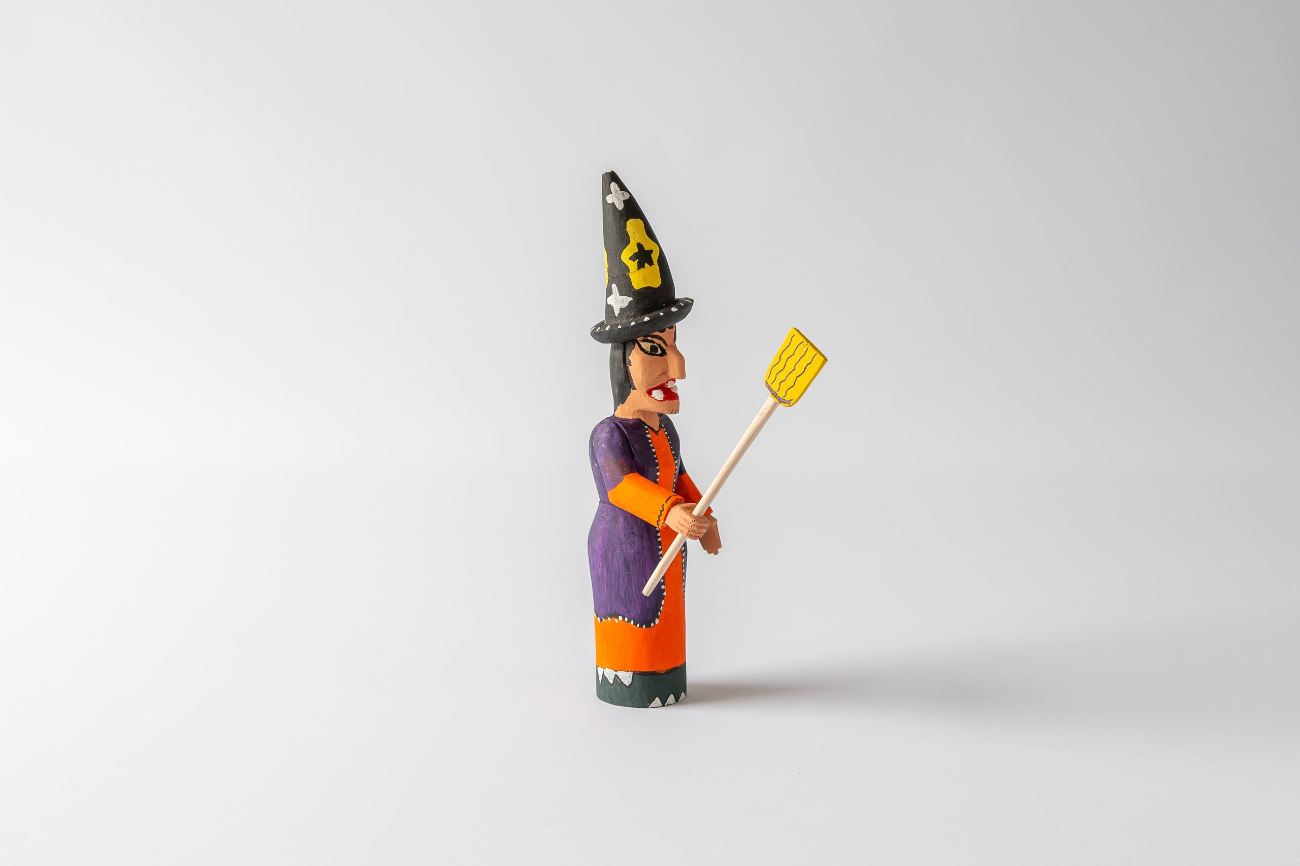 Woodcarving: witch with black high hat, orange dress with violet overcoat, broom